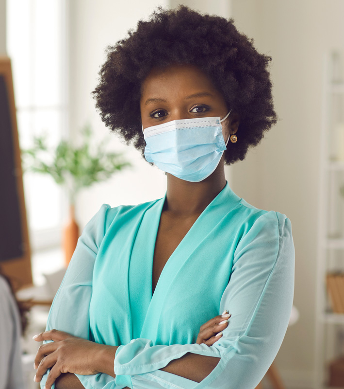 Portrait of black businesswoman with Afro hairstyle in medical face mask standing arms crossed in office. Young woman returns to work after Covid-19 lockdown ends. Coronavirus pandemic safety concept